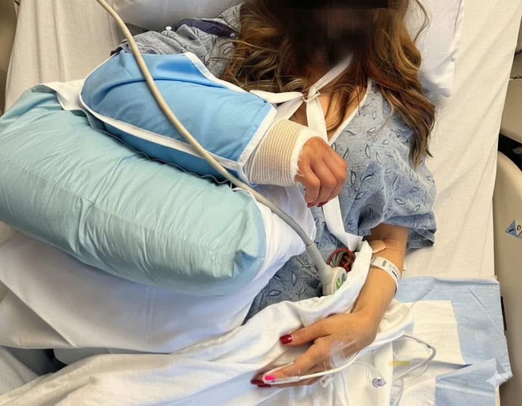 Maria in a hospital bed with a bulky post-surgical arm cast and IV lines following her wrist surgery from a car accident.