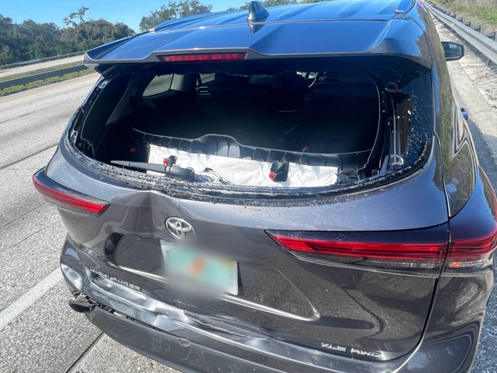 Rear view of a dark gray Toyota SUV on the highway shoulder with severe rear-end damage, shattered rear window, deployed interior airbags, and a blurred license plate.