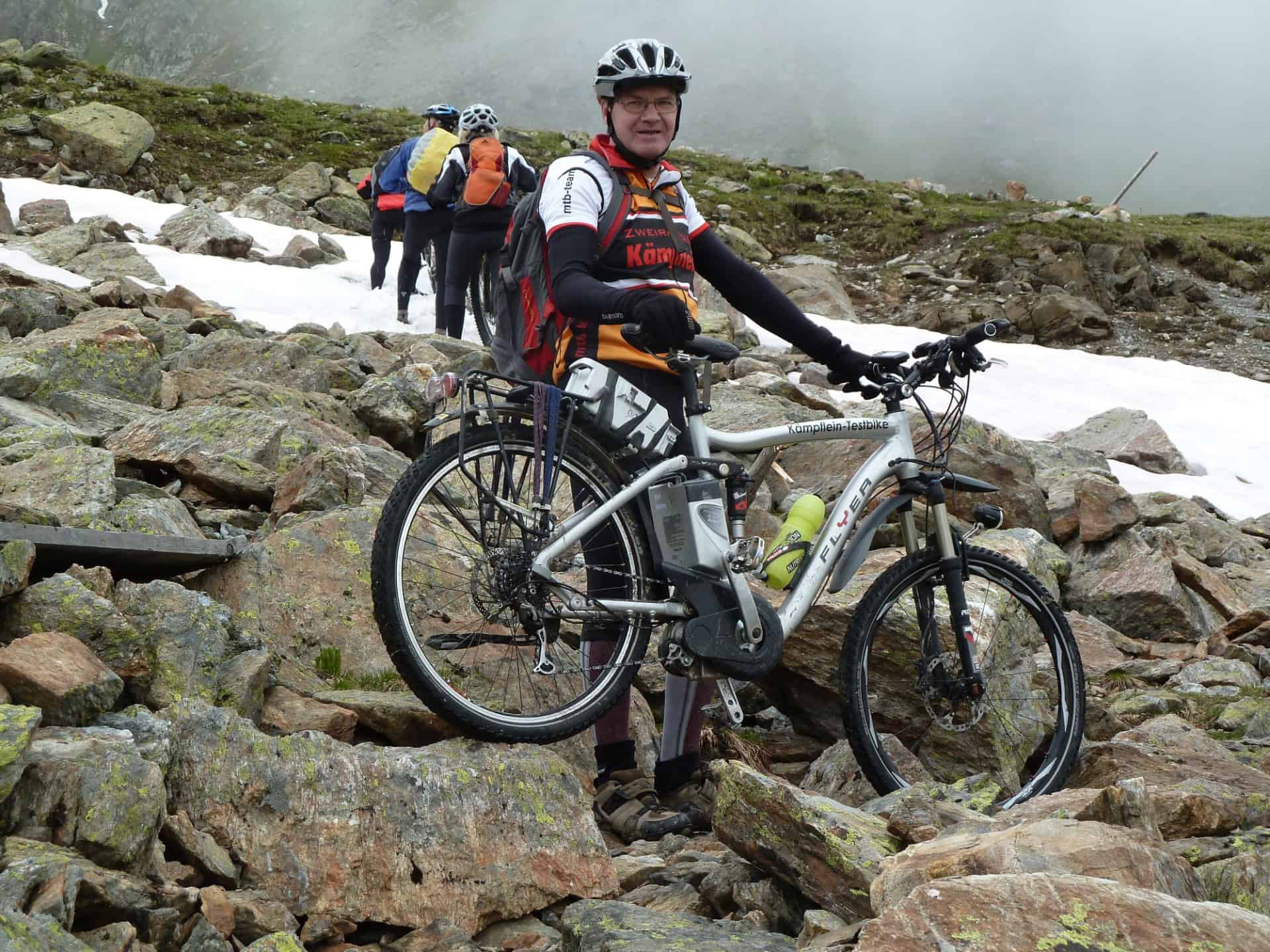 Mann mit Mountainbike auf felsigem Bergpfad bei Nebel und Schnee.