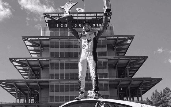 Race car driver celebrating victory on podium with trophy at Kellymoss event.