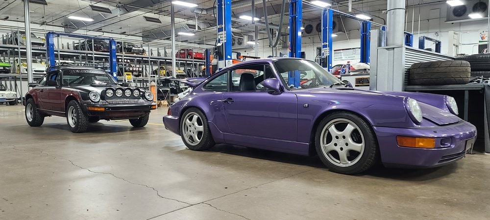 Purple Porsche 964 C4 in a workshop with classic cars in the background.
