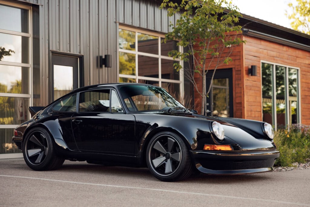 Black 1989 Porsche 964 parked outside modern building with trees.