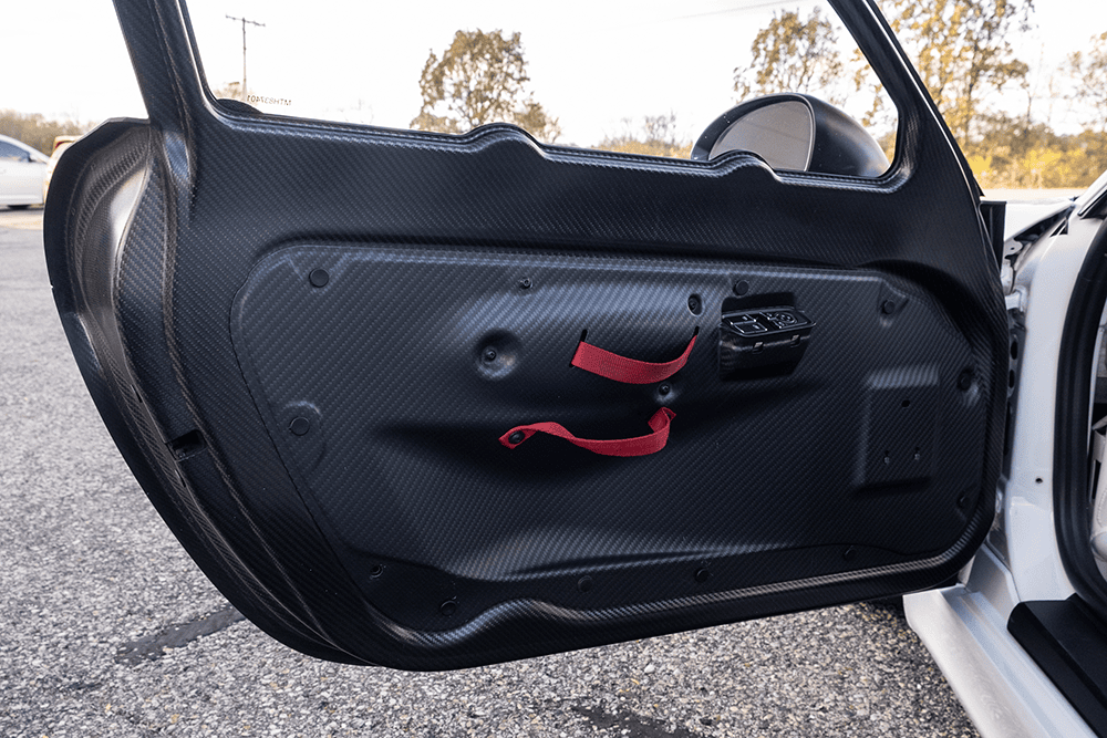 Close-up of a car door interior with carbon fiber panel and red straps.