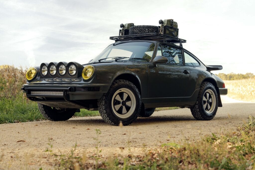 Classic black Porsche 964 with rally lights and roof rack in outdoor setting.