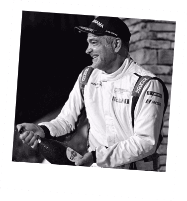 Race car driver smiling in racing suit and cap, holding a bottle of champagne.