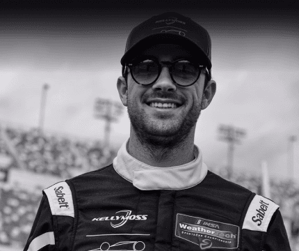 Race car driver smiling in racing suit at a track.