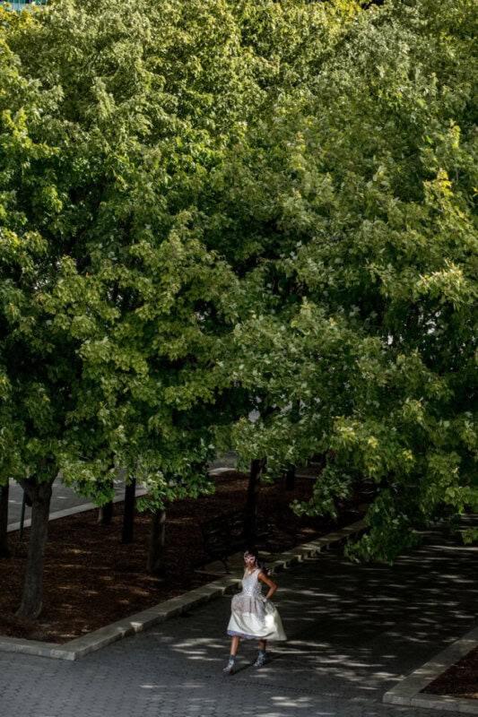 A model in an embellished white dress and decorative mask poses on a tree-lined urban walkway in dappled sunlight