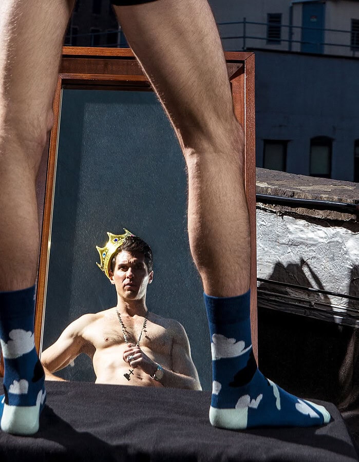 Male fashion model wearing a crown reflected in a rooftop mirror framed by another model’s legs.