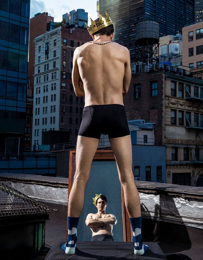 Man with a crown and shorts standing on rooftop with city skyline.