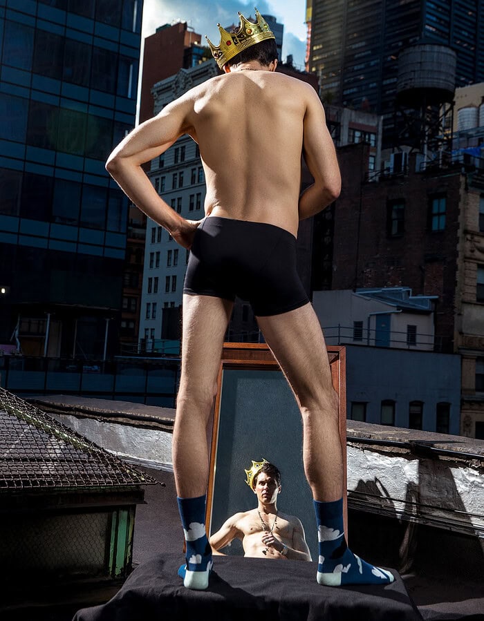 Young man with a crown looking at his reflection in a mirror on a city rooftop.