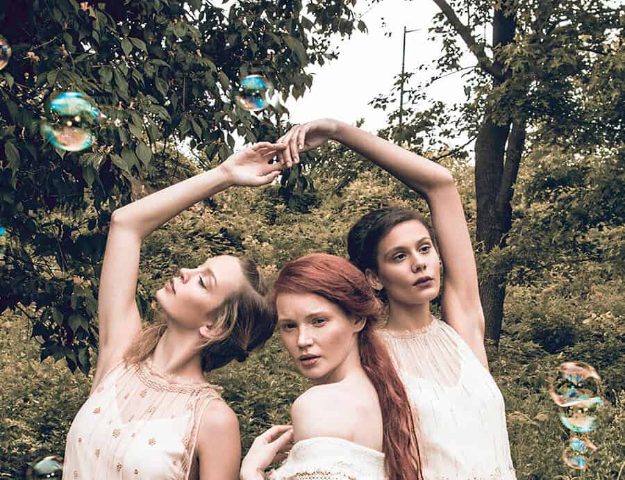 Three fashion models in pale dresses posing beneath raised arms with bubbles floating around them.