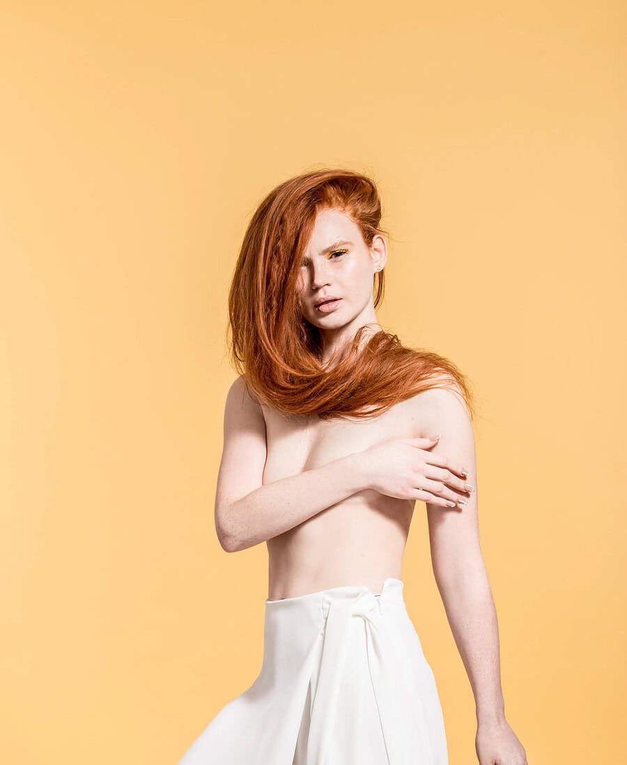 Red-haired model posing in white wide-leg trousers against a warm yellow studio background.