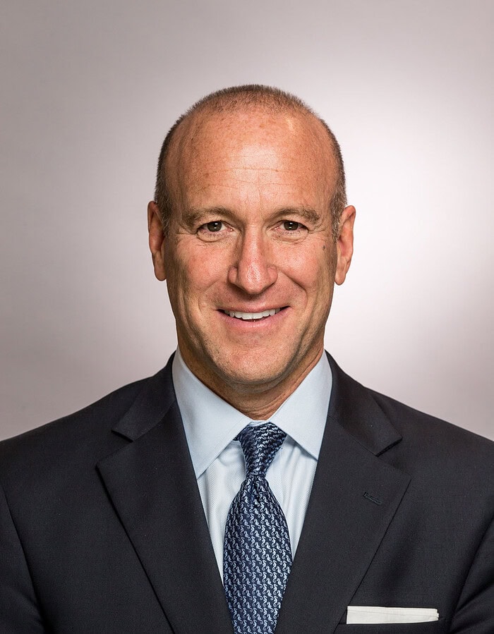 Male business executive in a dark suit and patterned tie photographed against a warm neutral background.