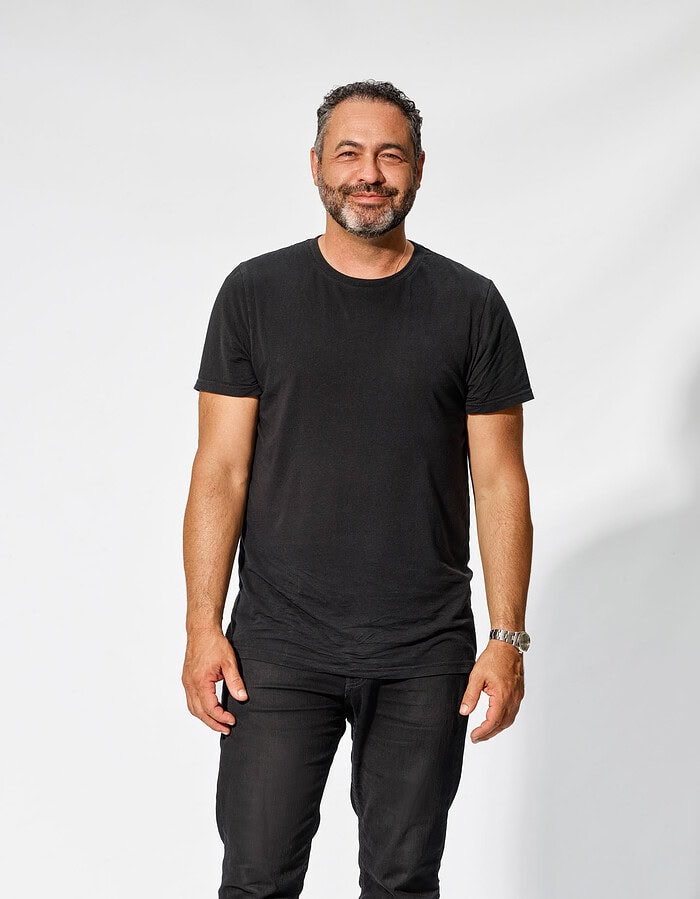 Male professional wearing a black T-shirt and dark pants against a white studio background.