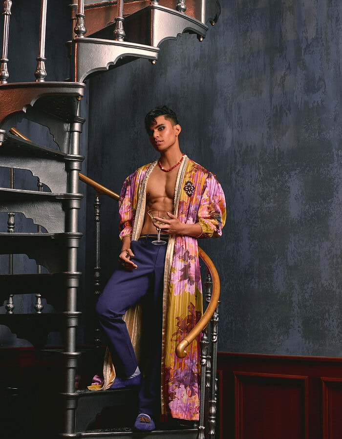 Menswear model in a long floral robe and purple trousers standing beside a cast-metal spiral staircase.