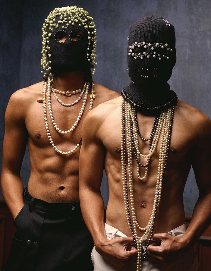 Two bare-chested models wearing black knitted face coverings decorated with pearls and jewelry.