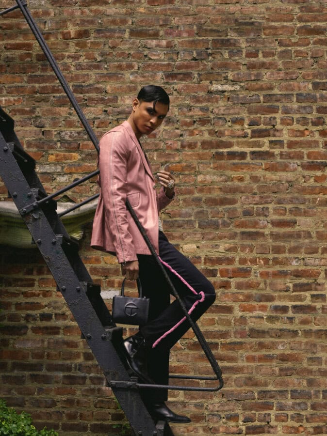 Model wearing a pink jacket, dark trousers and black boots while standing on an exterior fire escape.