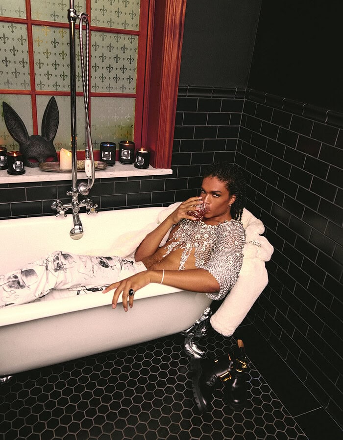 Wide view of a model reclining in a claw-foot bathtub surrounded by black tile, candles and decorative styling. Caption:
