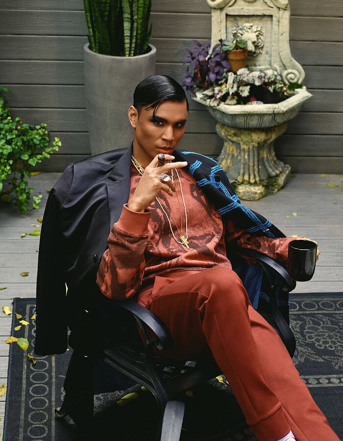 Model seated in a rust-colored fashion look with layered necklaces, rings and a dark coat.