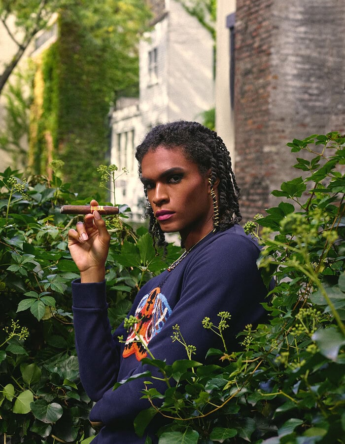 Model in a navy embroidered top holding a cigar while surrounded by dense green foliage.