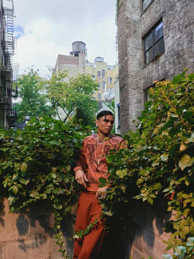 Model wearing a rust-colored outfit standing among ivy in an East Village courtyard.