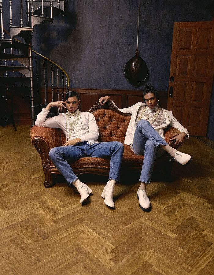 Two menswear models seated on an antique couch wearing white jackets, layered pearls and blue jeans.