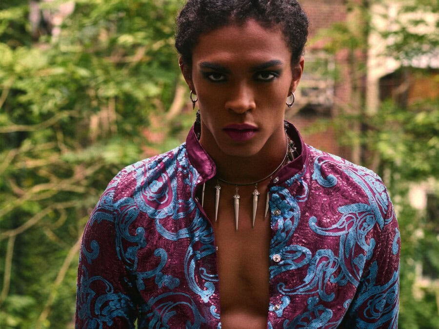 Close portrait of a menswear model wearing a burgundy and blue sequined jacket in a green courtyard.