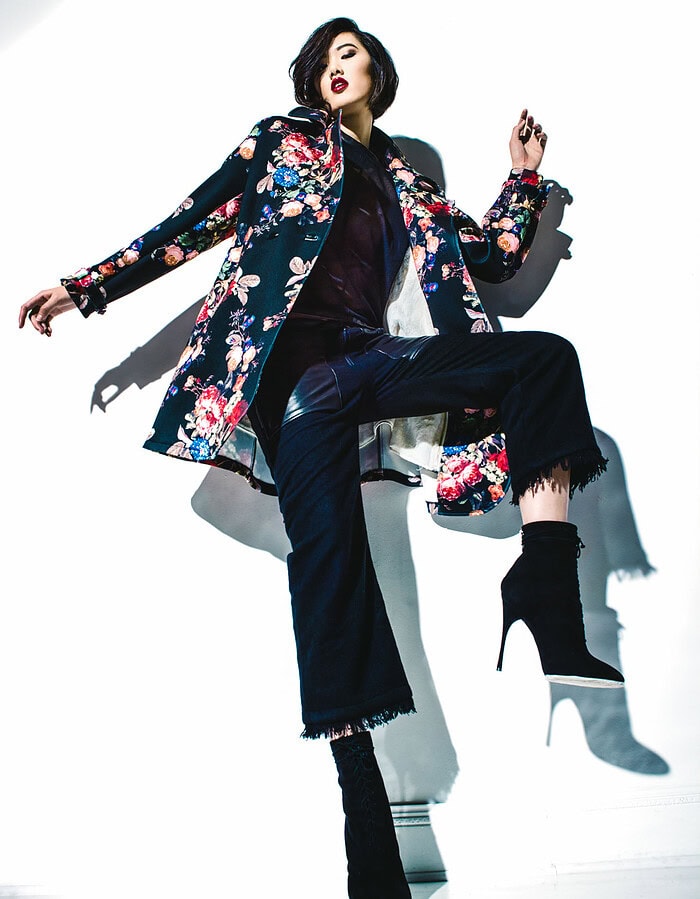 Model in a floral coat posing with one knee raised against a brightly lit studio wall.