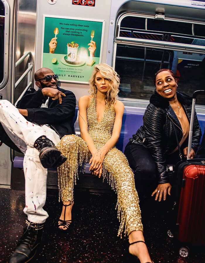 Fashion model in a gold outfit seated between two passengers inside a New York subway car.