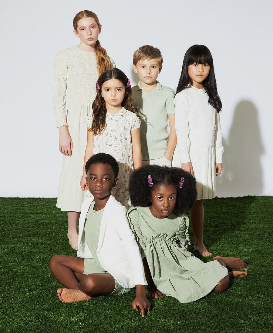 Six child models wearing cream and pale green outfits pose together on artificial grass.