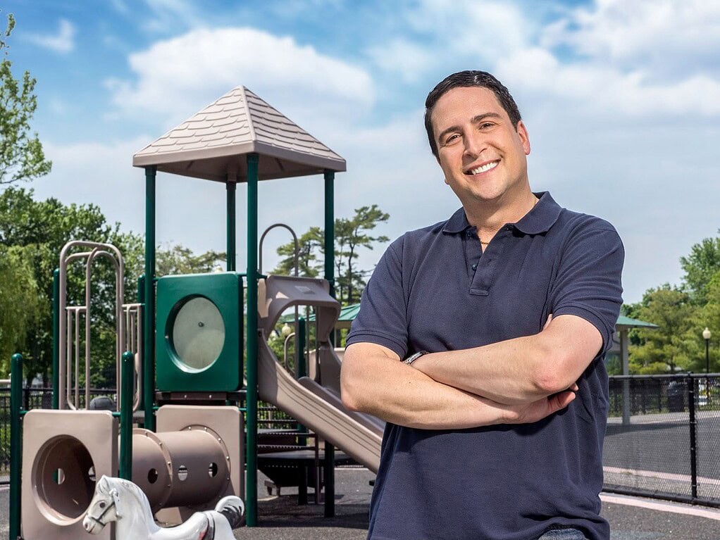Smiling man with folded arms photographed in front of a neighborhood playground.