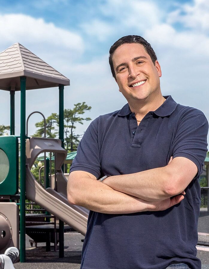 Smiling man with folded arms photographed in front of a neighborhood playground.