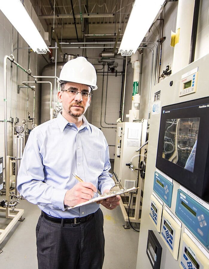 Man in a hard hat holding a clipboard inside a pharmaceutical production facility.