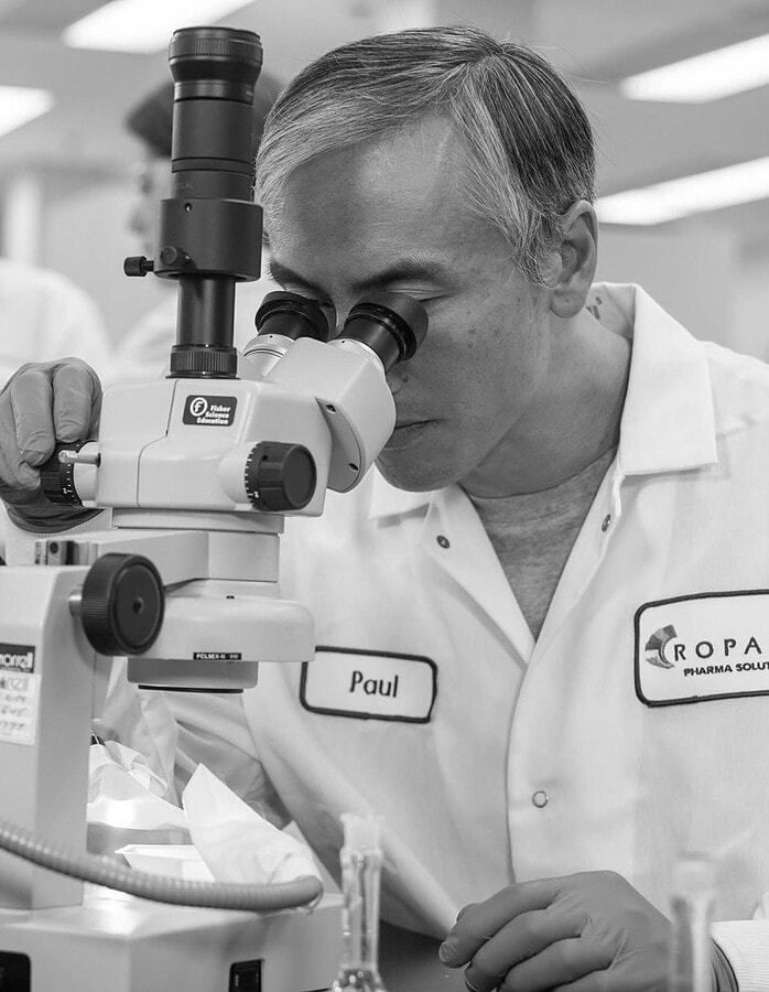 Scientist looking through a microscope in a pharmaceutical laboratory while colleagues work behind him.