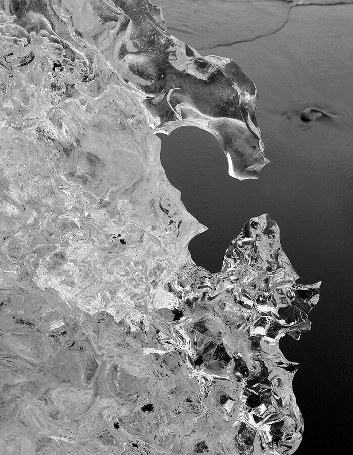Black and white close-up of jagged translucent ice beside dark water in Iceland.