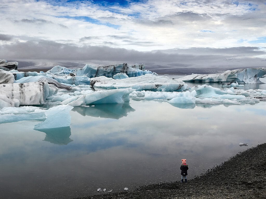 Small figure in a pink hat standing beside a quiet Iceland glacier lagoon with blue icebergs.