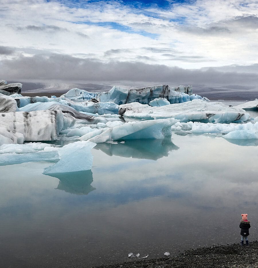 Small figure in a pink hat standing beside a quiet Iceland glacier lagoon with blue icebergs.