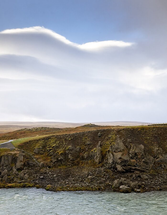 Wide Icelandic highland landscape with a river, cliffs, road, and pale dramatic clouds.