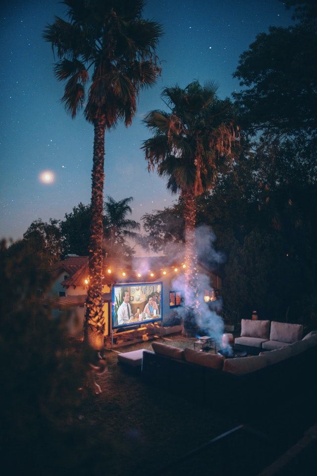 Outdoor movie night with palm trees, cosy seating, and a large screen under a starry sky.