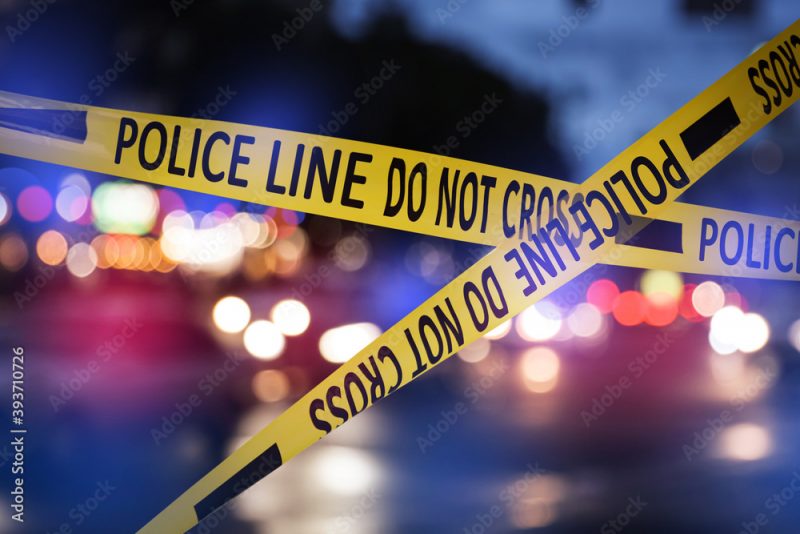 Yellow law enforcement tape isolating crime scene. Blurred view of city street, toned in red and blue police car lights