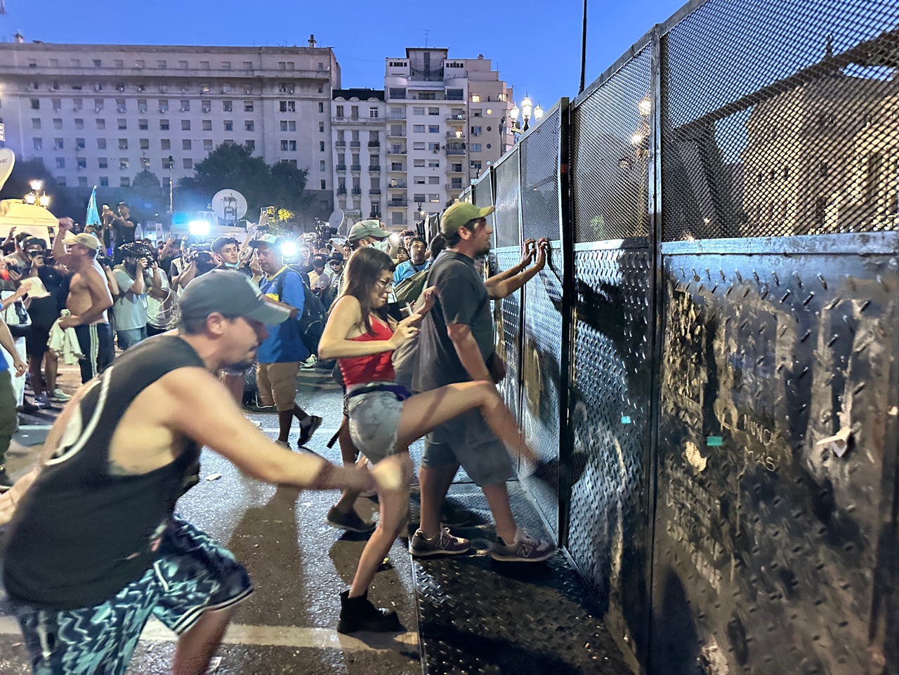 Protesters push and kick barricades outside Argentina’s National Congress as night falls, illuminated by streetlights and camera flashes.