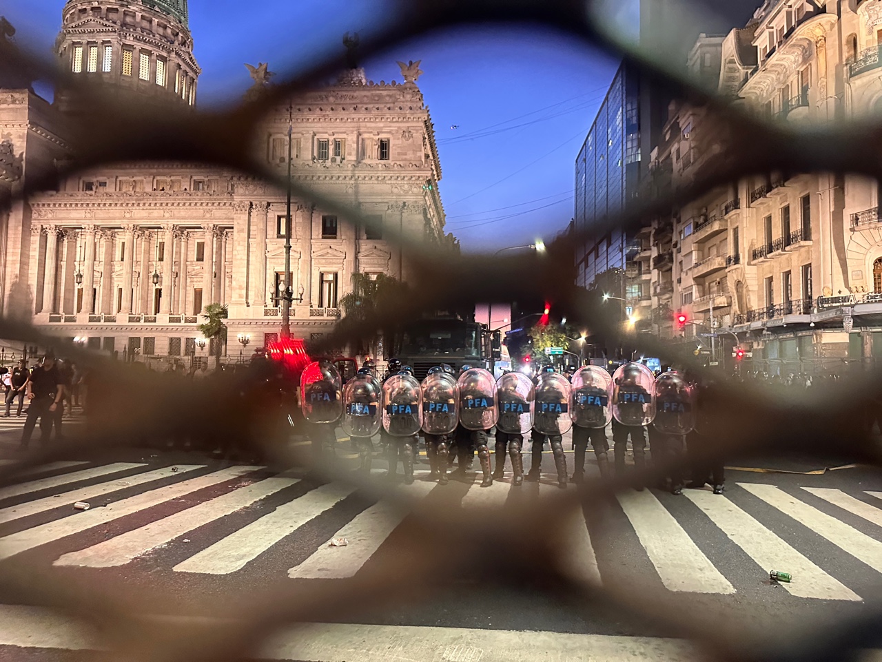 Federal Police officers stand behind barricades near Argentina’s National Congress, shields raised as water cannon trucks prepare for crowd control.