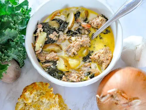 bowl of zuppa toscana next to bunch of kale, onion, garlic, and cheddar biscuit.