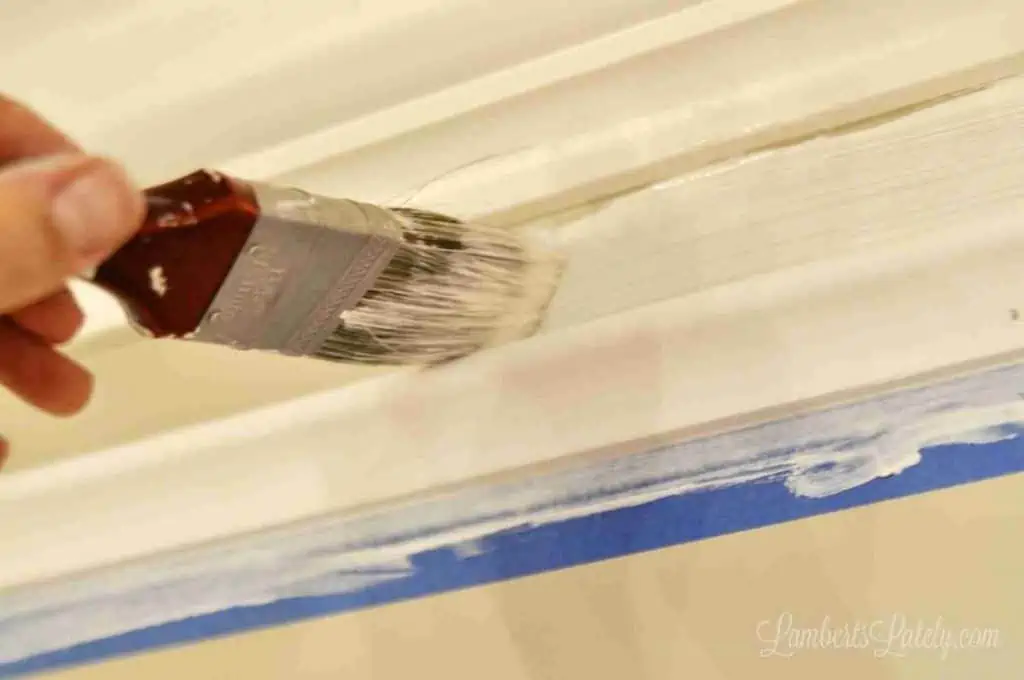 paint brush painting white paint in a gap between two pieces of crown molding on a wall.