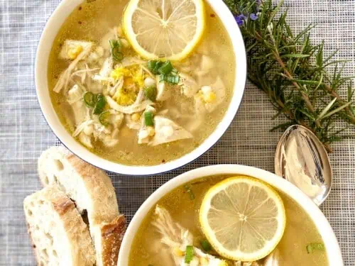 two bowls of greek chicken lemon soup, surrounded by bread and herbs.