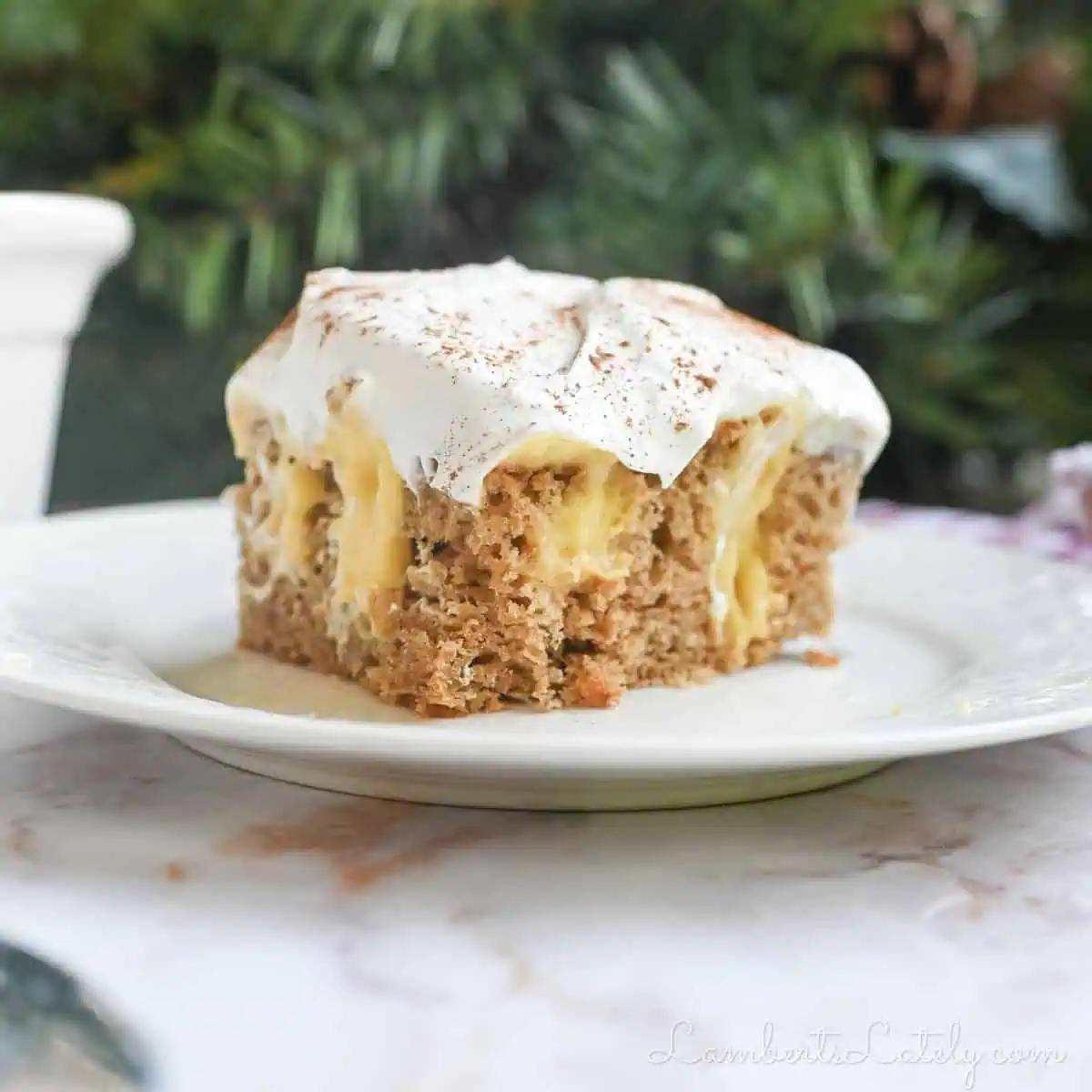 piece of eggnog poke cake on a plate in front of greenery and a baking pan.