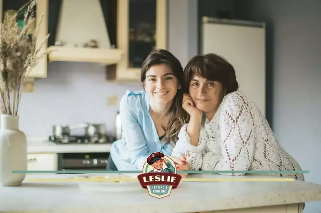 Two women leaning on a kitchen counter, happily posing for a photo. At the bottom center, the logo for "Leslie Heating & Cooling" proudly stands, reflecting their commitment to excellent customer service.