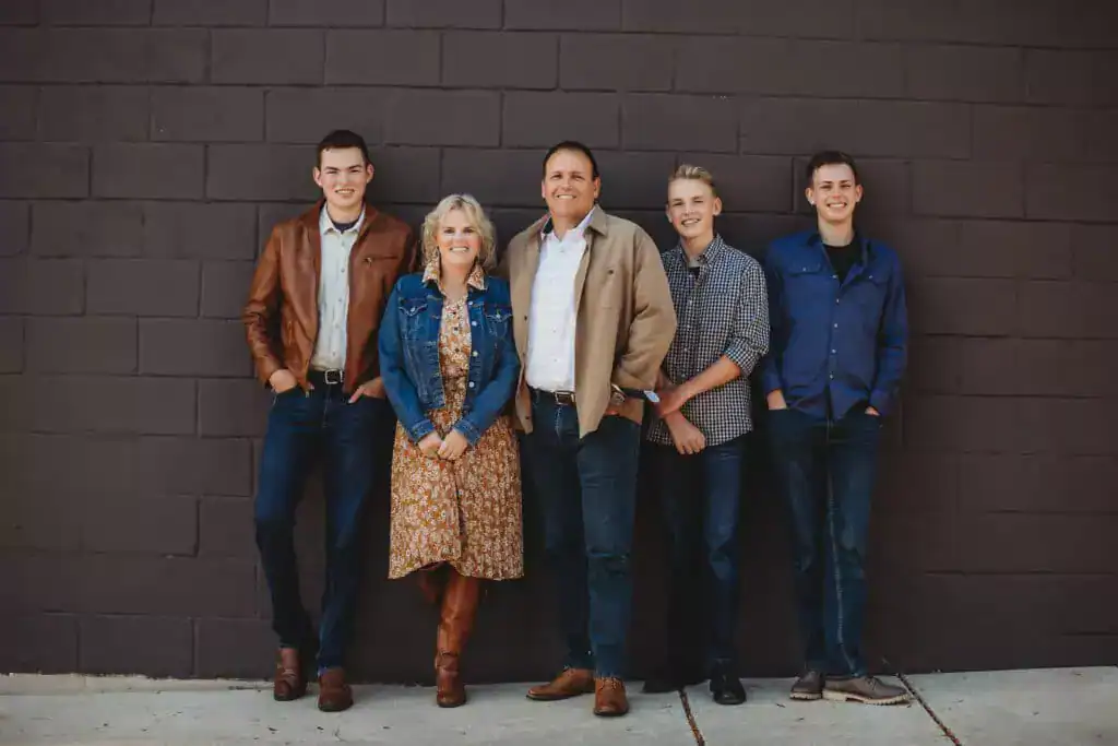 A group of five people stands in front of a brick wall. They are wearing casual clothing and all are smiling at the camera.