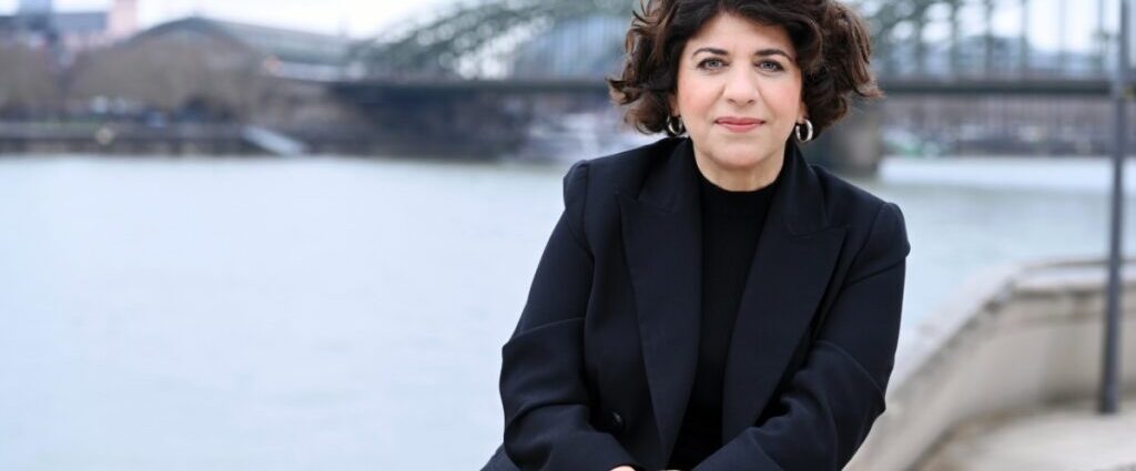 Lindweiler Frau am Flussufer in einer schwarzen Blazer, mit Brücke im Hintergrund, urbane Szenerie, entspannte Atmosphäre, professionelle Ausstrahlung, modern gekleidet, stilvoll, Business-Umfeld, städtischer Kontext, elegant.