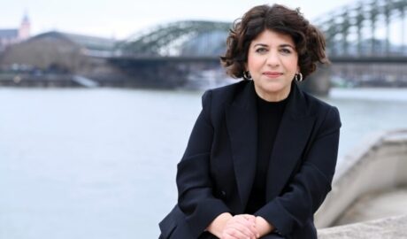 Lindweiler Frau am Flussufer in einer schwarzen Blazer, mit Brücke im Hintergrund, urbane Szenerie, entspannte Atmosphäre, professionelle Ausstrahlung, modern gekleidet, stilvoll, Business-Umfeld, städtischer Kontext, elegant.